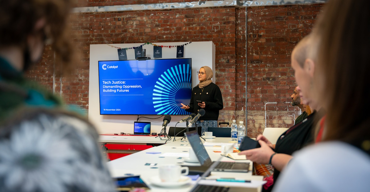 A photo of Sheeza Shah at the previous in-person Funders’ Forum. Sheeza is a Brown woman wearing glasses and a hijab. She is standing next to a screen with the words “Tech justice: dismantling oppression, building futures”. The profile of a white woman and Brown man are visible looking on. 