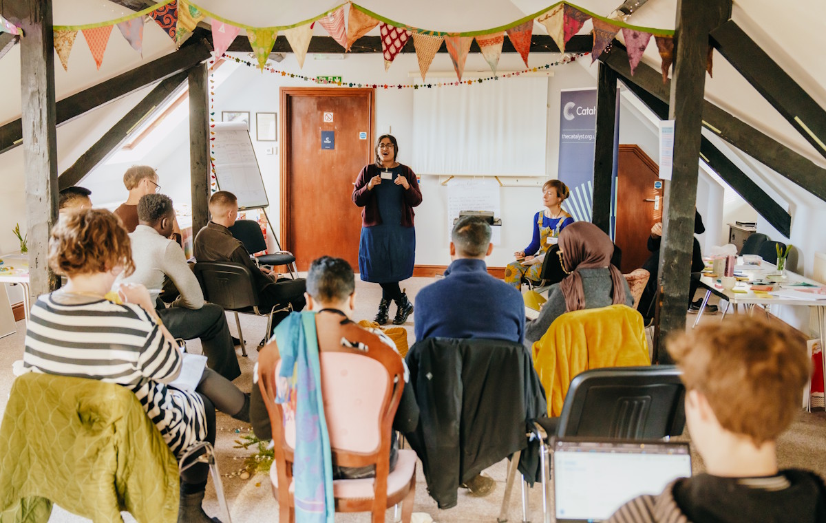 Sadaf, a brown skinned woman with glasses, speaks to the group from the front of the room at the Kindling retreat while others sit in a circle listening.