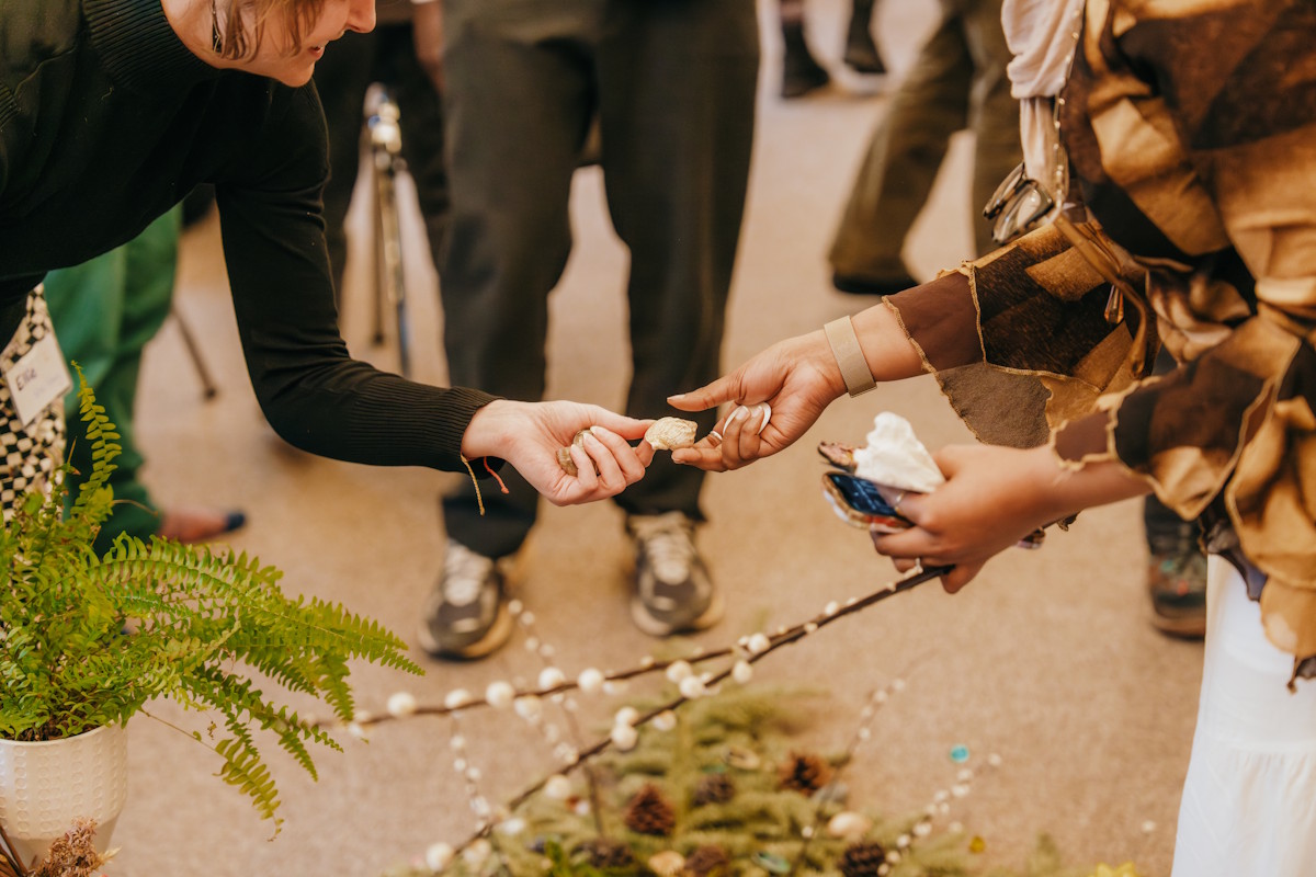 Close-up of two people’s hands exchanging small natural objects over a floor arrangement of branches and ferns during a retreat ritual.